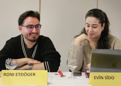 Roni und Evîn vom Vorstand sitzen an einem Tisch. Sie lächeln, schauen aber nicht direkt in die Kamera. Die Schilder stehen vor ihnen.