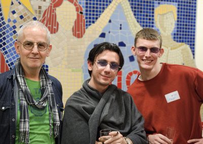 Drei Männer stehen nebeneinander vor einem Mosaik in blau / rot. Alle drei tragen eine Brille und lächeln in die Kamera